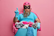 Suprised overweight superhero in helmet tsirt and rubber gloves stands with basin of dirty linen and detergents indicates above poses against pink background. Peope house chores and cleaning concept