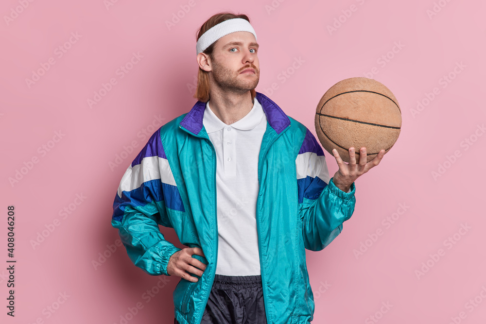 Serious man basketball player holds ball looks confidently at camera wears white headband sportsclothes enjoys playing favorite game isolated over pink background. Sport and recreation concept - obrazy, fototapety, plakaty 