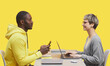 Side view portrait of two contemporary business people sitting opposite each other at desk during meting and smiling against yellow background