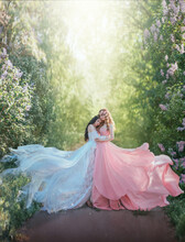 Fantasy Photo Two Young Beautiful Glamorous Women Stand Hugging In Spring Garden On Path. Woman Princess In White Silk Vintage Dress Fabric Flying In Wind. Girl Queen In Pink Gown. Green Nature Trees