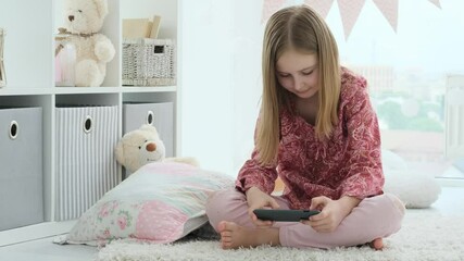 Sticker - Smiling little girl playing game on smartphone sitting on floor in bright room