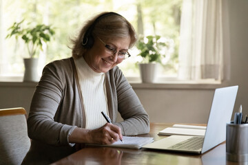 Smiling mature woman wearing headphones writing notes, using laptop, sitting at table at home, aged female wearing glasses listening to lecture, watching webinar, studying online, learning language