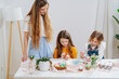 © zzzdim - two daughters painting easter eggs with brushes behind a table at home and young mother watching them