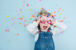 © zzzdim - Little girl in happy birthday glasses making funny face, lies on a blue-surfaced floor with colorful confetti pieces. Top view.