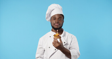 Portrait of African american chef cook in uniform showing dessert cupcake standing isolated over blue background. Bakery or happy birthday concept