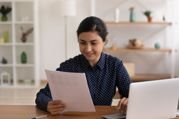 Canvas Print - Confident indian female small business owner sit by desk with laptop study paper letter of supplier choose samples of production to order buy. Young hindu woman freelancer doing paperwork at workplace