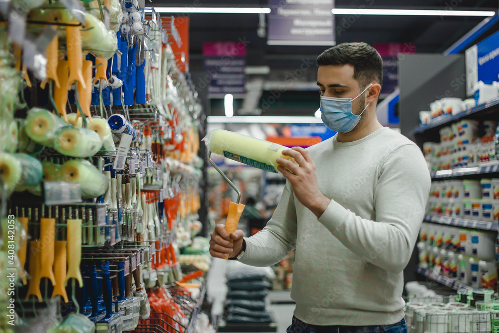 A buyer wearing medical mask chooses a paint roller in a hardware store. - obrazy, fototapety, plakaty 