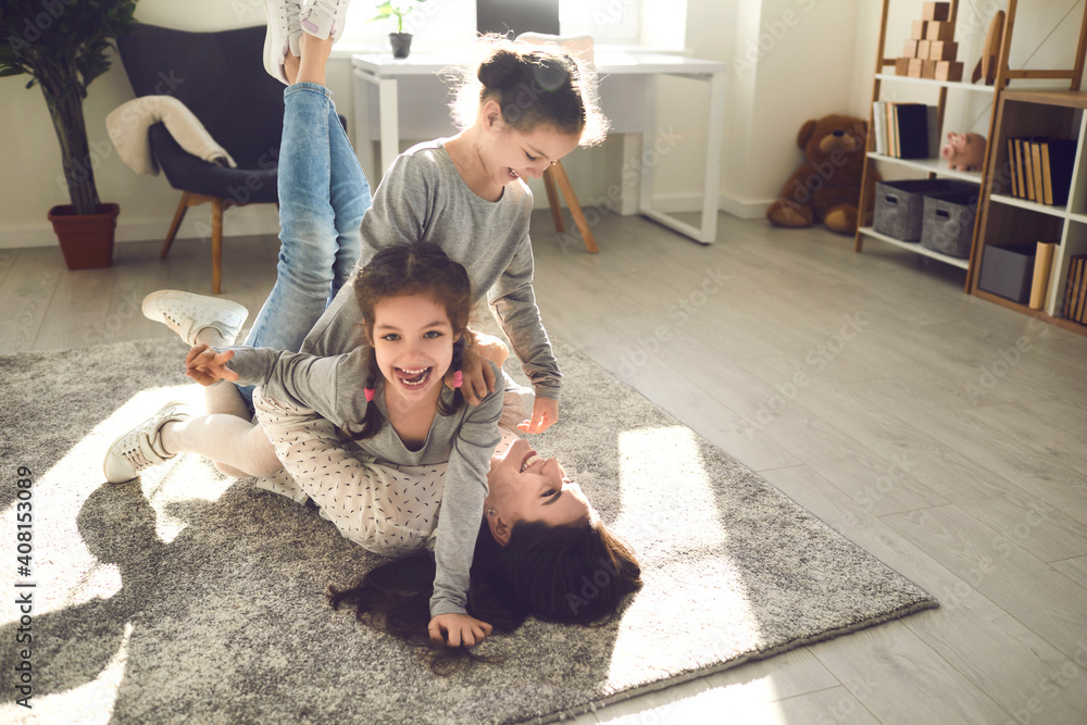 Family enjoying quality time and having fun at home. Young mom and little kids playing on carpet backlit with sunlight. Happy mother and cute carefree children fooling around and laughing on floor rug - obrazy, fototapety, plakaty 