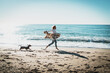 Smiling blonde woman running alongside the sea side with her pet dog on a sunny day. Cheerful Caucasian female in blue jeans with plaid on her shoulders playing with her doggy at the sandy beachside.