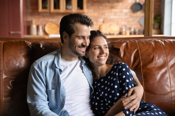 Wall Mural - Happy young Caucasian man and woman sit rest on sofa at home hug and cuddle look in distance dreaming thinking. Smiling couple relax on couch embrace imagine or visualize good future together.