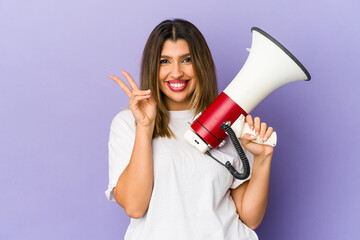 Wall Mural - Young indian woman holding a megaphone isolated joyful and carefree showing a peace symbol with fingers.