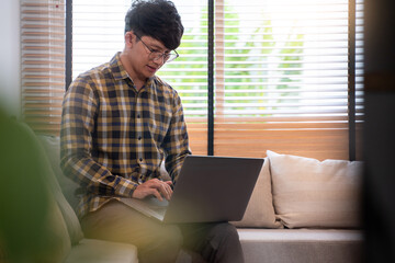 young attractive asian man relaxing comfortably working on laptop in bright living room at his home.