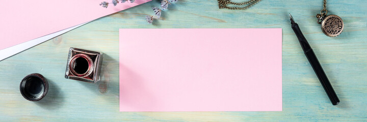 Wall Mural - Stationery panorma with a pink greeting card or invitation, overhead flat lay shot with ink on a blue background