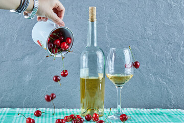 Woman hand throwing cup of cherries and a bottle of white wine with glass on blue background