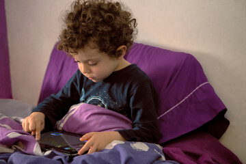 4-5 year old boy with curly hair lying down watching cartoons on tablet, Happy boy lying in bed playing in the morning, Kid having fun and relaxing alone in the room, New normal lifestyle