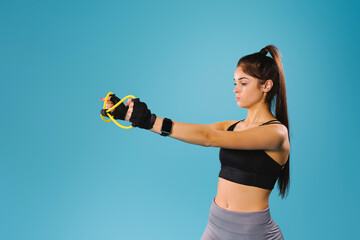 A focused athletic girl in a black top stretches a rubber band in front of her to train her arms against a blue background with side space.