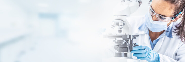 female lab technician in protective glasses, gloves and face mask sits next to a microscope and coni