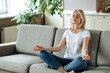 © Kateryna - Satisfied attractive middle aged female sits on sofa at home and chilling. Blonde mature is practise yoga and meditation in lotus position with eyes closed, relieve stress, calmness concept