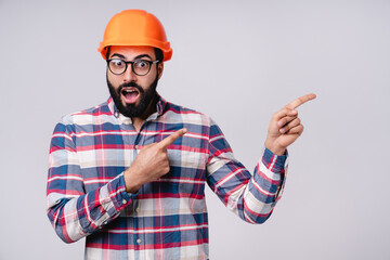 Wall Mural - Surprised Arabian construction worker in orange hardhat pointing at copy space isolated over grey background