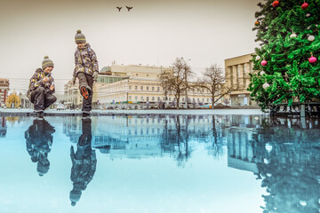 Two boys walking in the winter in the city square look at their reflections in a huge puddle. Around the city architecture, the sky, birds and a Christmas tree decorated for the New Year