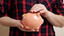 Young Woman In Black Red Checkered Shirt Holds Small Pink Piggy Bank On Palm And Throws Bronze Coin Inside Against Peach Wall Closeup