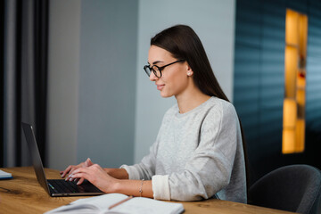 Happy young woman in eyeglasses smiling while working with laptop