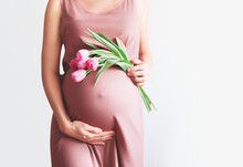 Beautiful Pregnant Woman With Tulips Flowers Holds Hands On Belly. Young Woman In Dress Waiting For Baby Birth. Pregnancy, Motherhood, Mother's Day Holiday Concept.