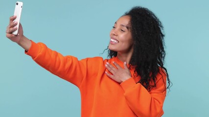 Wall Mural - Fun cute young african american woman with curly hair lush hairstyle 20s years old wearing orange sweatshirt doing selfie shot on mobile phone isolated on pastel blue color background studio portrait