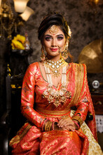 Magnificent Young Indian Bride In Luxurious Bridal Costume With Makeup And Heavy Jewellery Is Sitting In A Chair In With Classic Vintage Interior In Studio Lighting. Wedding Fashion.