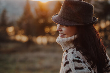 Wall Mural - Portrait of a woman in a large warm comfy sweater wearing a hat, smiling to the side, with sunset over trees and lake in the background