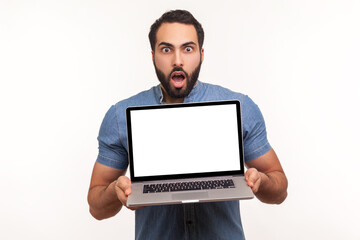 Shocked bearded man showing laptop with empty display, surprised with advertising area, looking at camera with big eyes and opened mouth