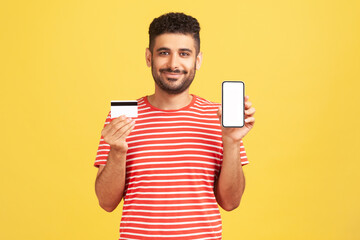 Cheerful positive bearded man in striped t-shirt holding and showing empty display smartphone and credit card, satisfied with easy mobile banking
