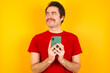 © Roquillo - Young Caucasian man wearing red t-shirt standing against yellow background holds telephone hands reads good youth news look empty space advert