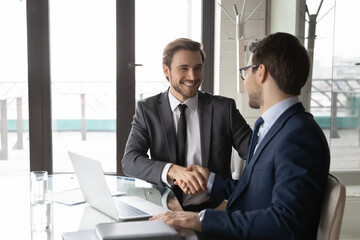 Wall Mural - Smiling businessman shaking client hand, closing successful deal, sitting at table with laptop in office, satisfied hr manager hiring new employee, business partners handshaking at meeting