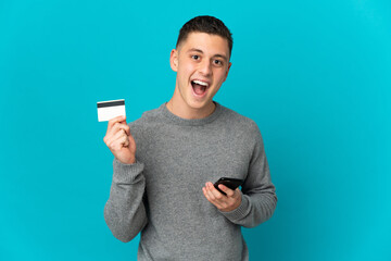 Young caucasian man isolated on blue background buying with the mobile and holding a credit card with surprised expression