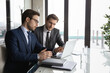 © fizkes - Confident businessman pointing at laptop screen, training new employee, sitting at table in modern office with panoramic windows, serious mentor coach teaching worker, colleagues working on project