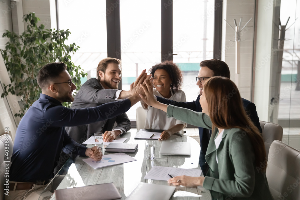 Excited diverse business team giving high five at briefing, sitting at table in boardroom, motivated for shared success, overjoyed colleagues joining hands, engaged in team building activity - obrazy, fototapety, plakaty 