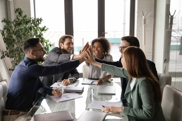excited diverse business team giving high five at briefing, sitting at table in boardroom, motivated