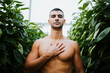 © Miguel Frias/Westend61 - Handsome shirtless man standing with hand on chest amidst plants at greenhouse
