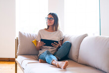 Thoughtful Brunette Businesswoman Sitting With Digital Tablet On Sofa In Living Room At Home
