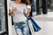 © zadorozhna - Woman using phone and walking near mall with coffee after shopping.