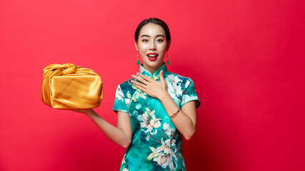 Wall Mural - Surprised Asian woman wearing traditional qipao dress holding golden gift box in isolated red studio background for Chinese new year concepts