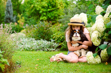 happy kid girl relaxing with her cavalier king charles spaniel dog in summer garden, sitting on lawn and having fun