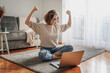 Pretty curly happy young woman sitting on floor at home in front of laptop with victory gesture with hands, successful and satisfied