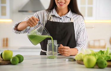 Wall Mural - Young woman pouring fresh green juice into mason jar at table in kitchen, closeup