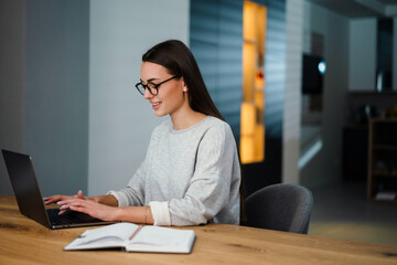 Happy young woman in eyeglasses smiling while working with laptop