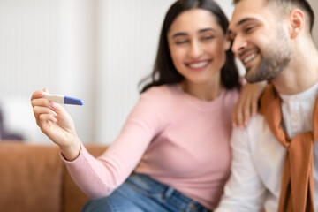 Wall Mural - Joyful Arab Couple Hugging Celebrating Positive Pregnancy Test Sitting Indoor