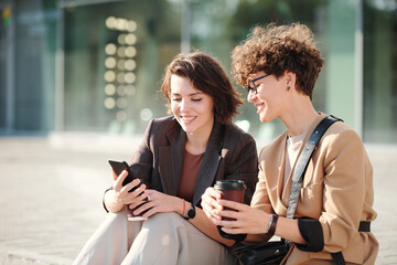 Canvas Print - Two young pretty female colleagues with drinks looking at screen of smartphone