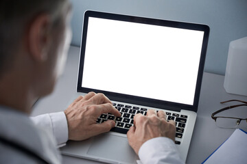 Male doctor using laptop computer with blank white mockup screen technology for tele medicine medical healthcare tech website ad concept, video call e telehealth online appointment. Over shoulder view