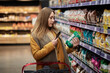 © Evgenia - A young woman scans the product with an app in her smartphone. To find out information about the product using new technologies. There is a large shopping cart nearby.